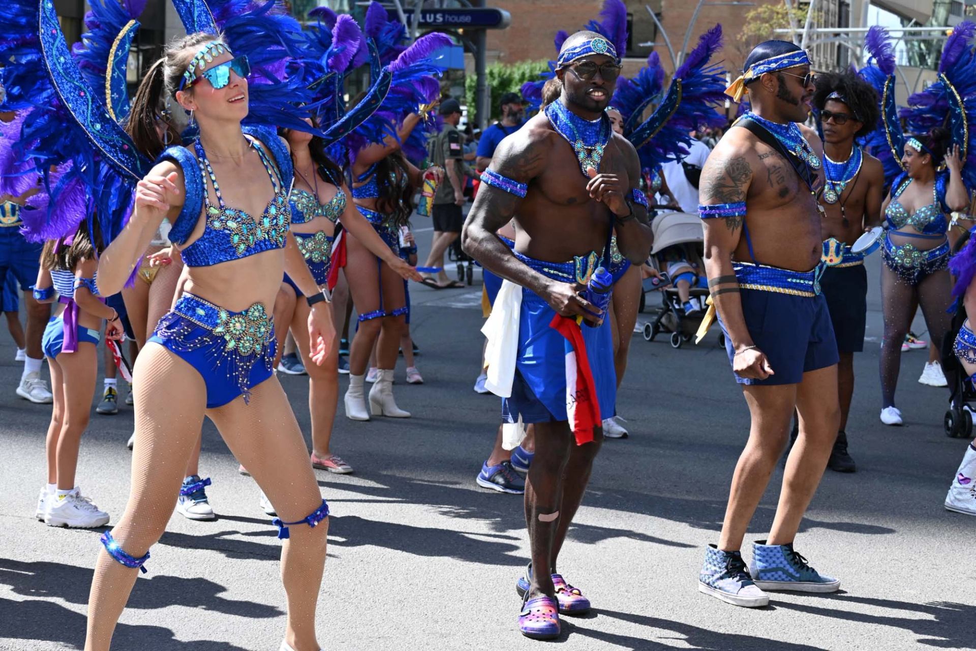 Parade performers in blue feathered costumes dance along a city street with spectators nearby.