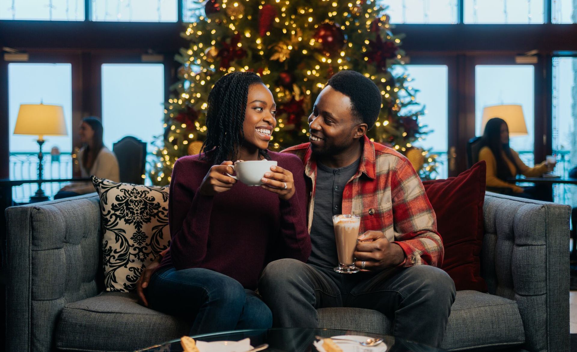 Two people enjoying hot drinks in a cozy room with a lit Christmas tree behind them.