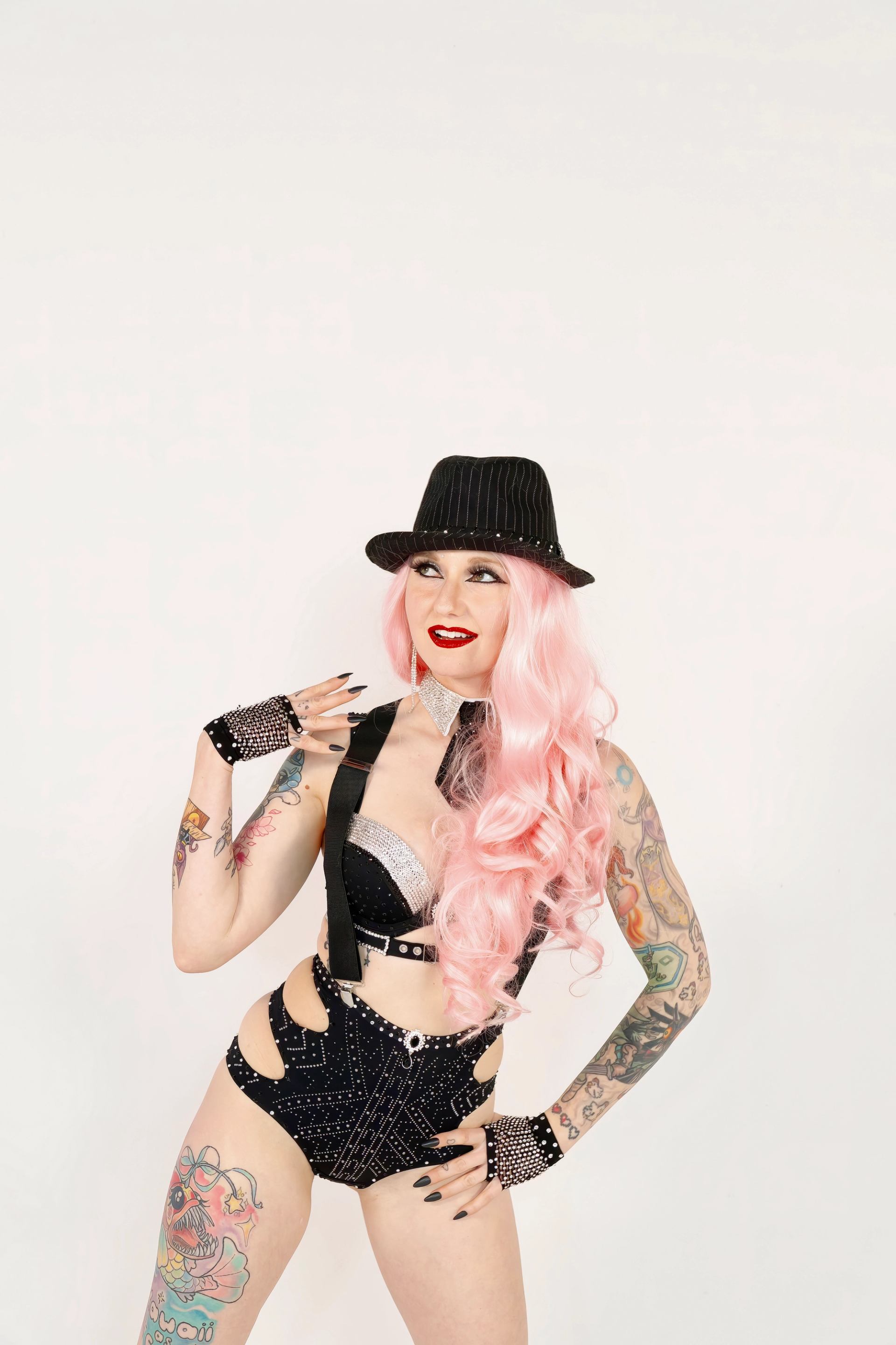 Woman with pink hair and tattoos wearing a black sparkly burlesque outfit and fedora, looking up.