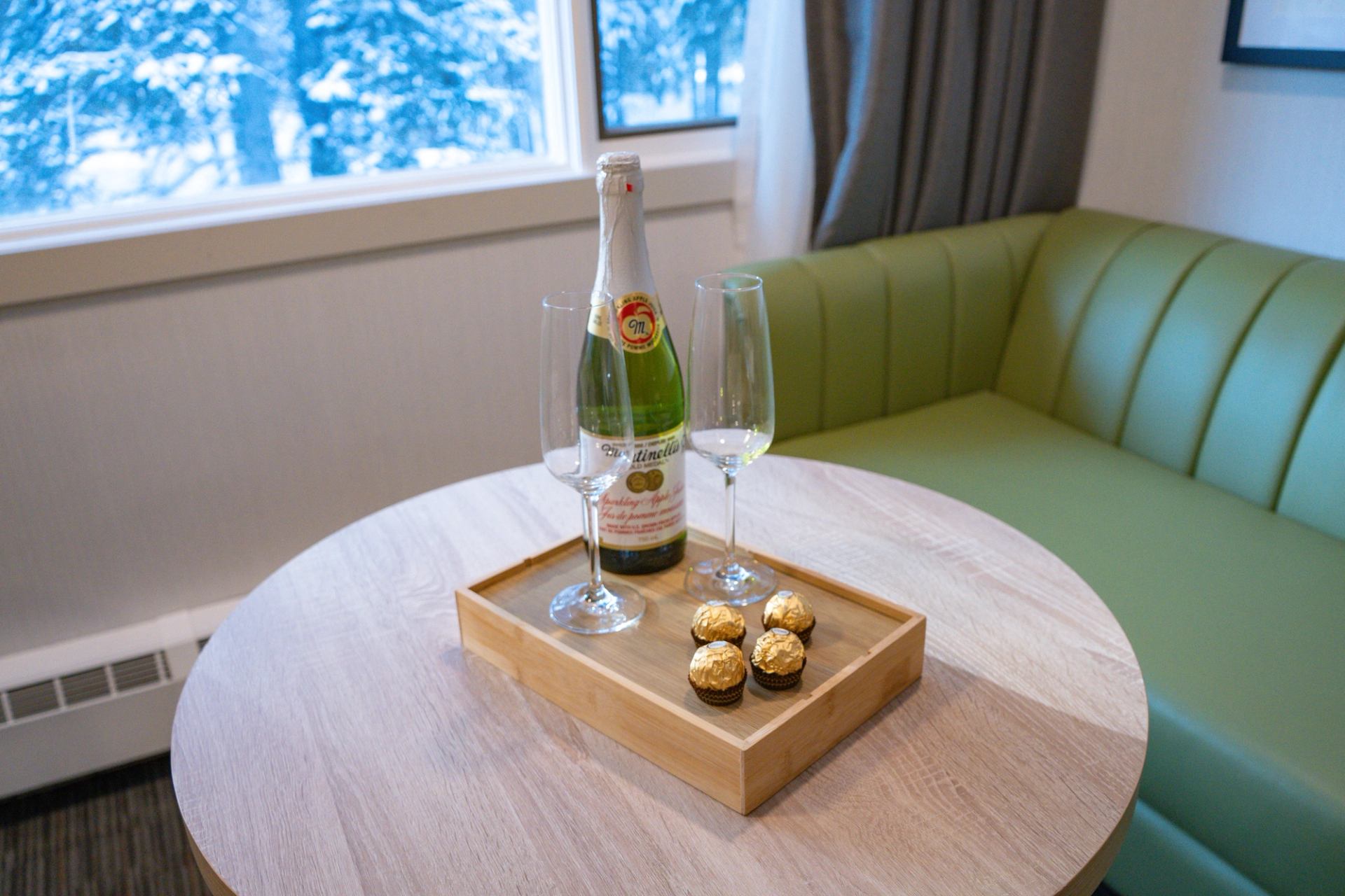 Tray with sparkling cider, two glasses, and chocolates on a table in a lodge room with snowy views.