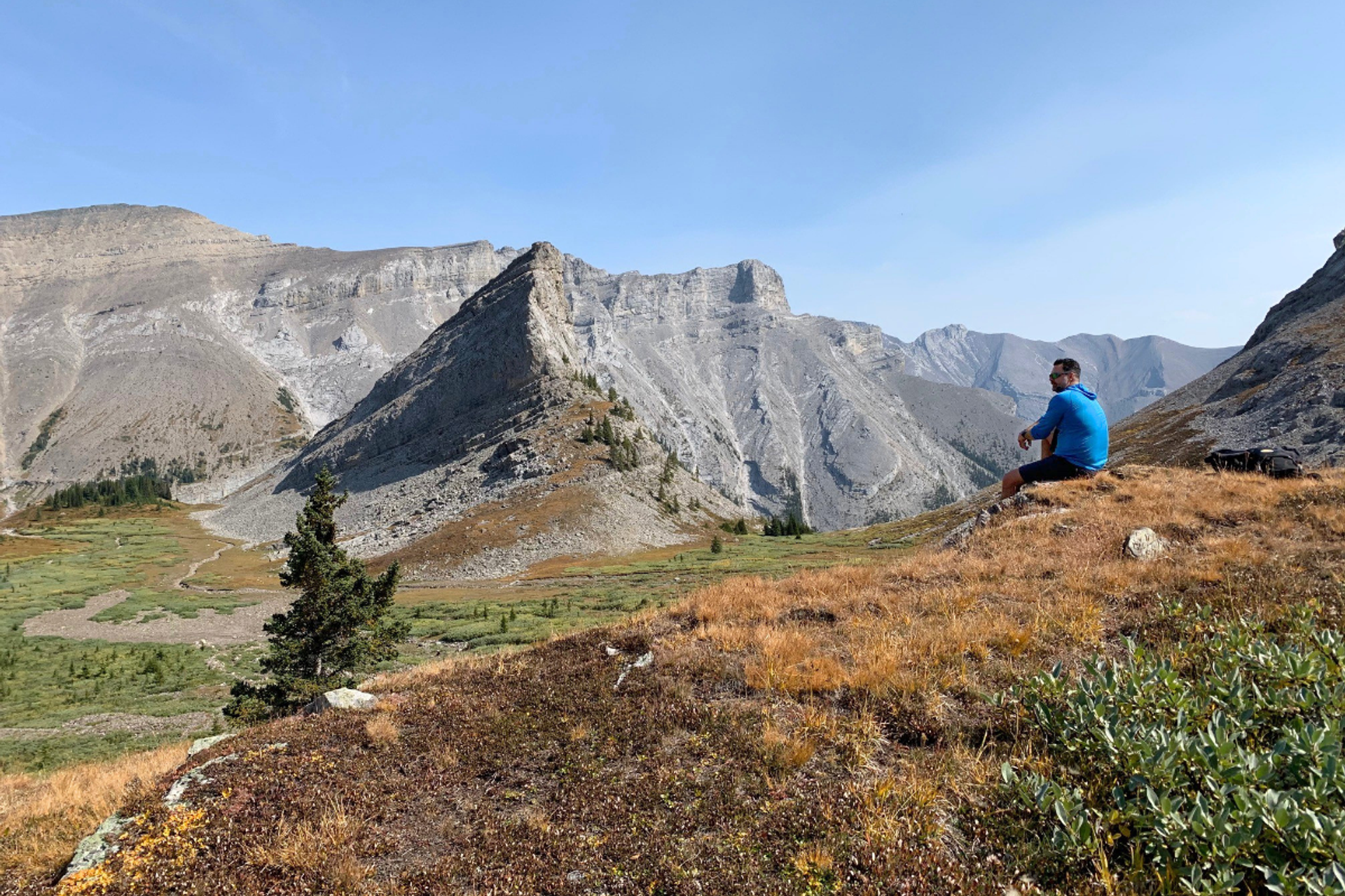 Hiker sitting on a ridgeline overlooking a wide alpine valley and rugged mountain peaks.
