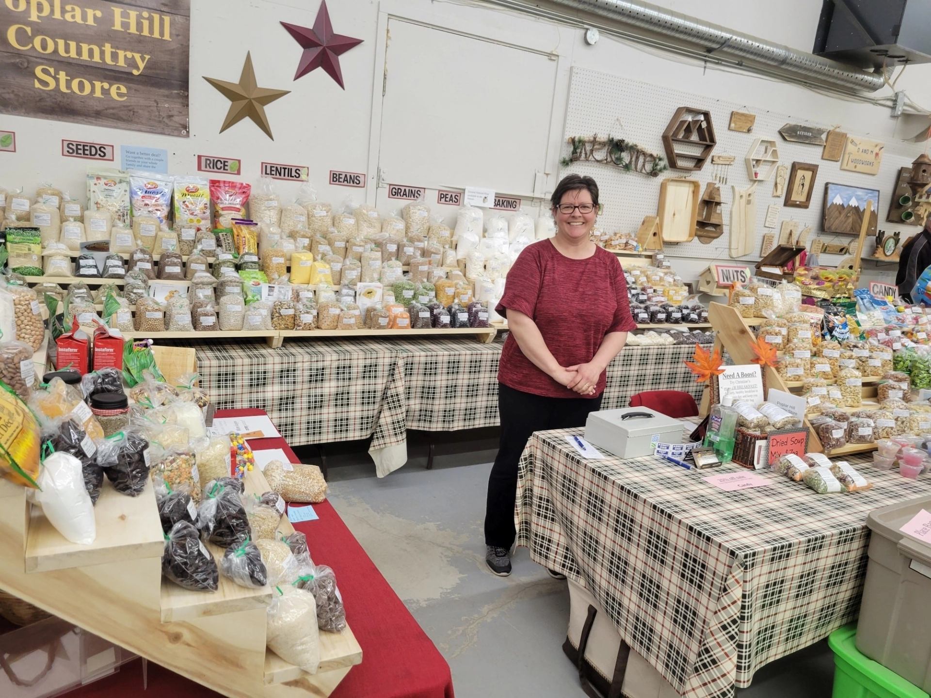 Market stall with bags of grains, seeds, mixes, and homemade goods displayed on plaid‑covered tables.