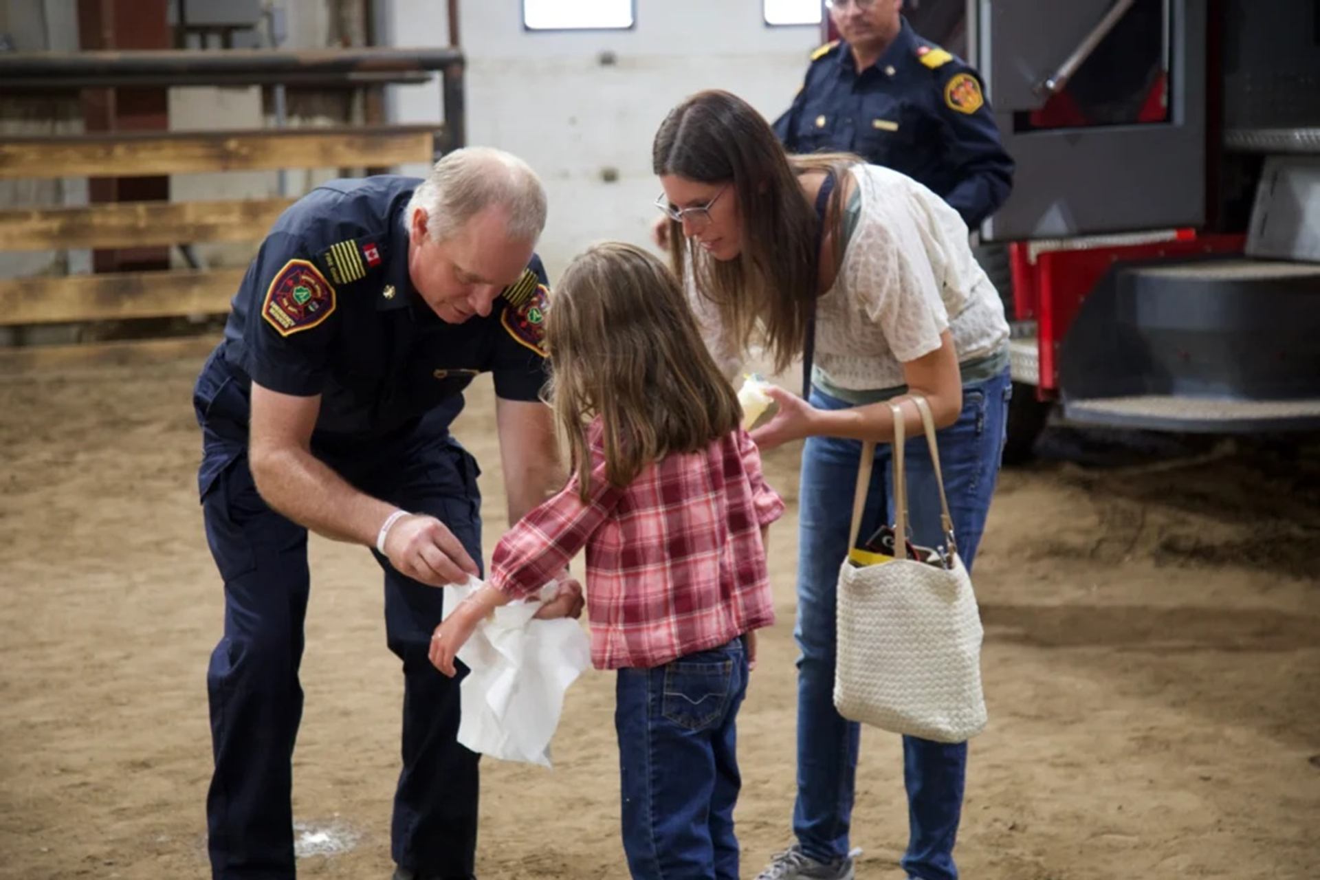 Firefighter kneels to assist child in plaid shirt while adult stands nearby indoors.