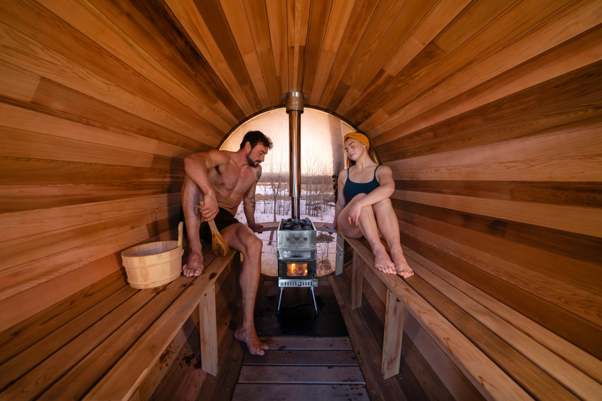 Two people sit on wooden sauna benches beside a small metal wood-burning stove.