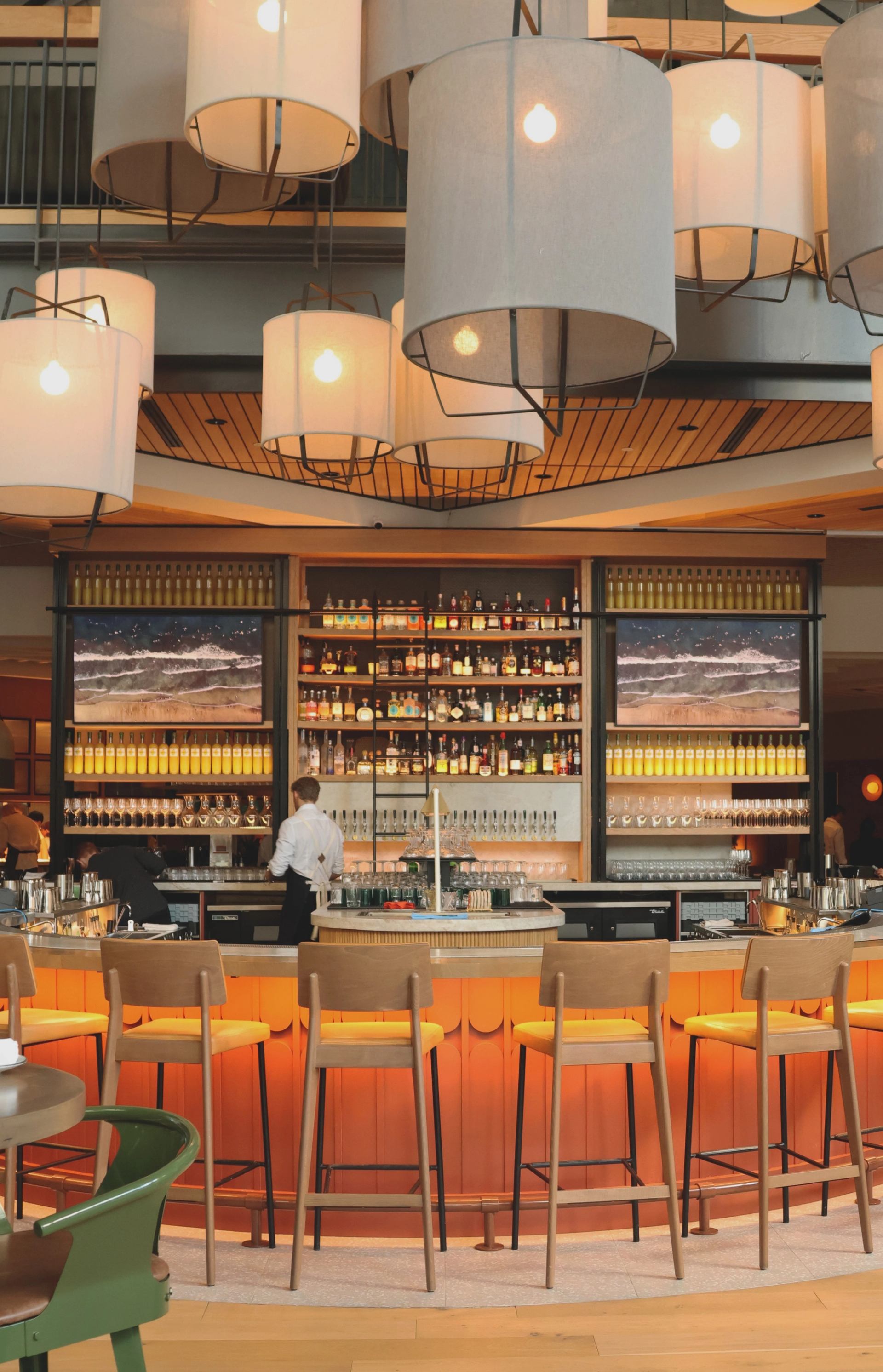 Bar counter with illuminated bottles, orange chairs, and hanging pendant lamps in restaurant interio.