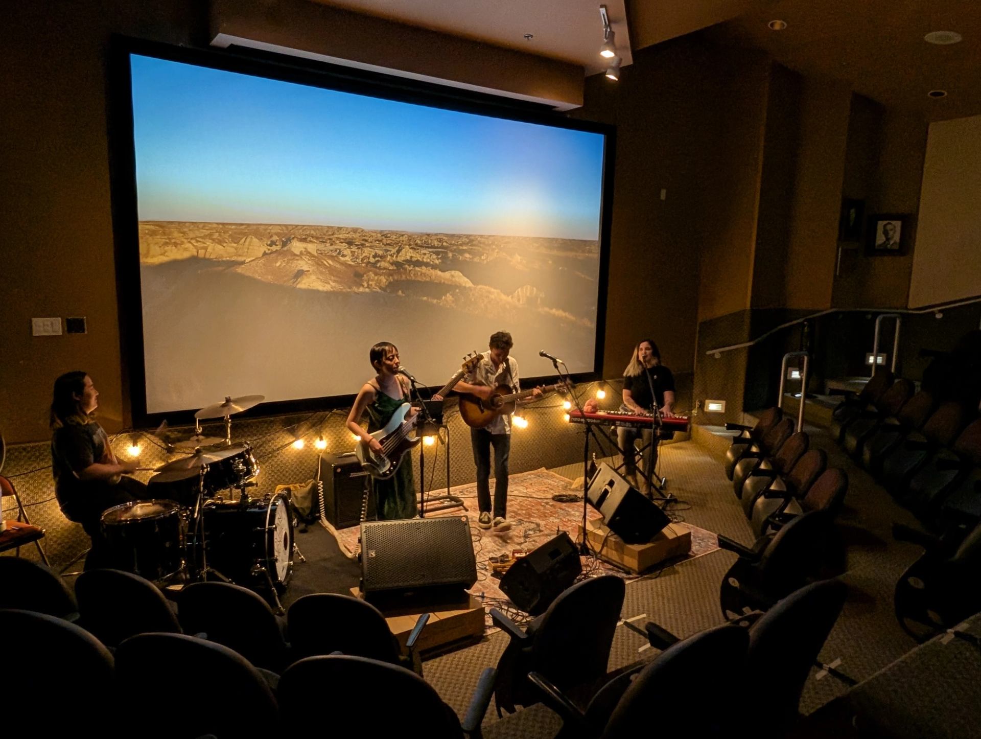 Band performs with drums, guitars and keyboard before a desert landscape screen.