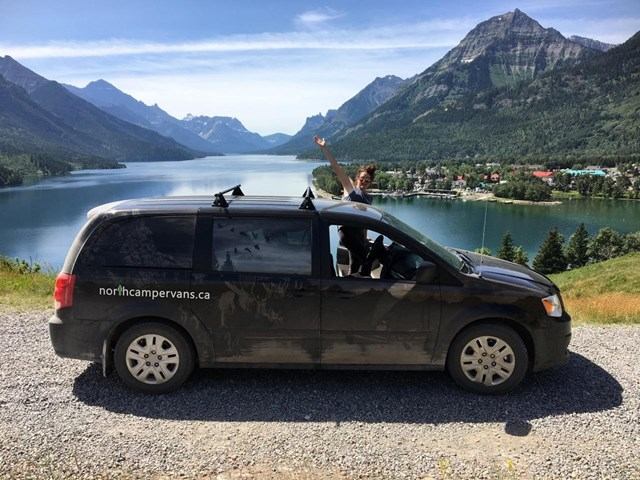 Black campervan parked on gravel road with lake, mountains, and town in the background.