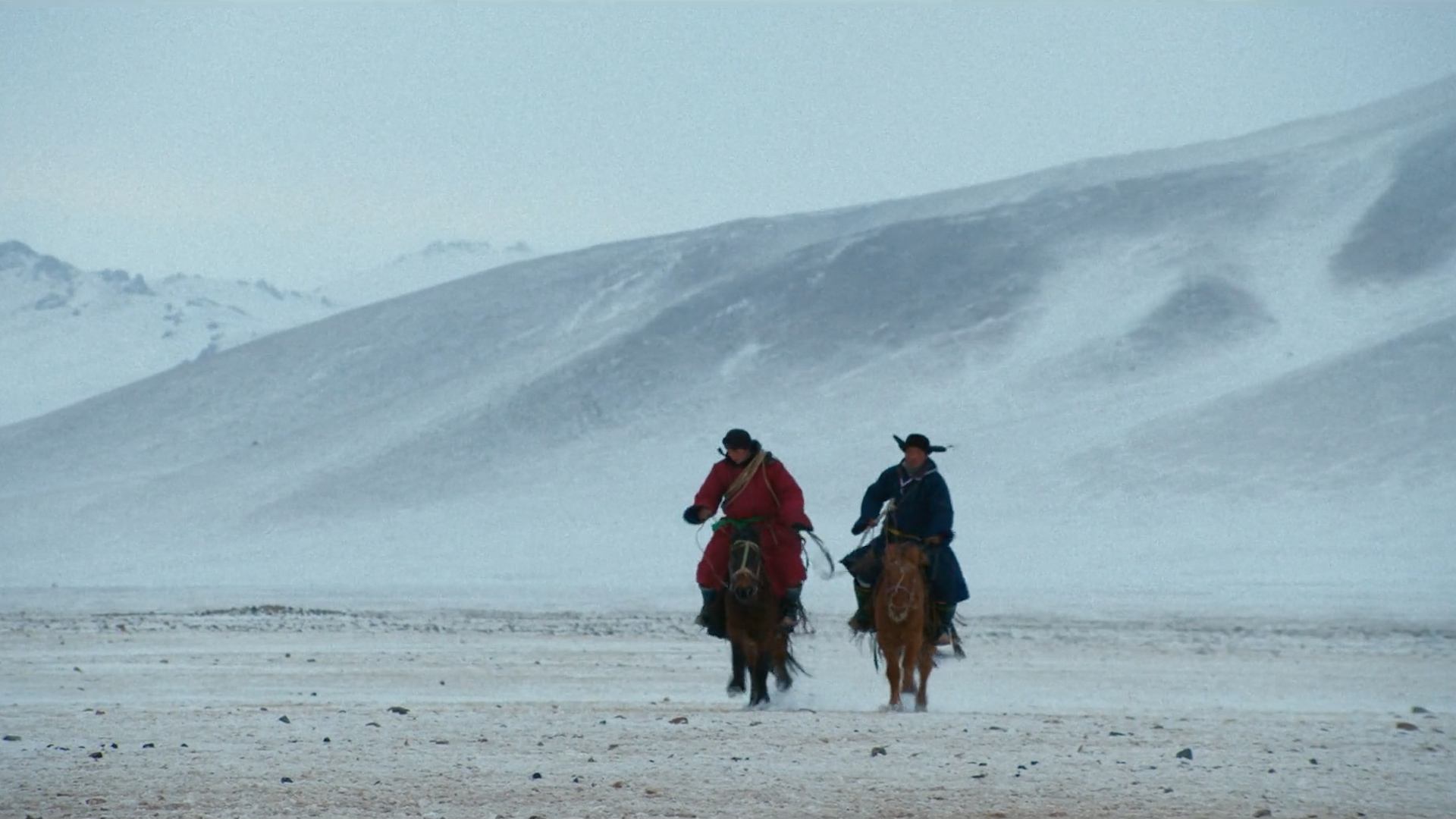 Two riders on horseback crossing a vast, wind-swept winter plain with snow-covered hills behind.