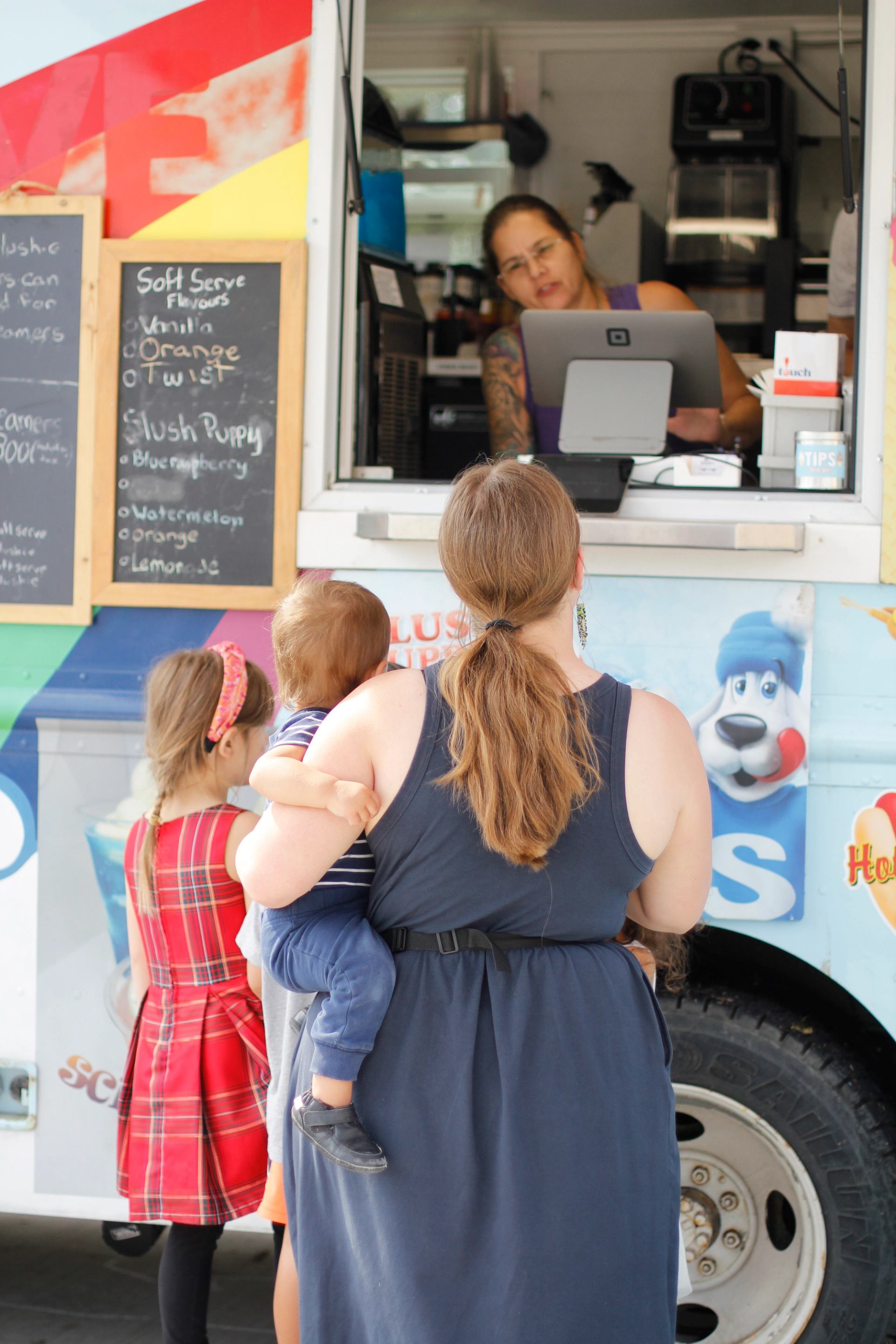 Adults and children line up at a colourful food truck with a menu board during an outdoor market event.