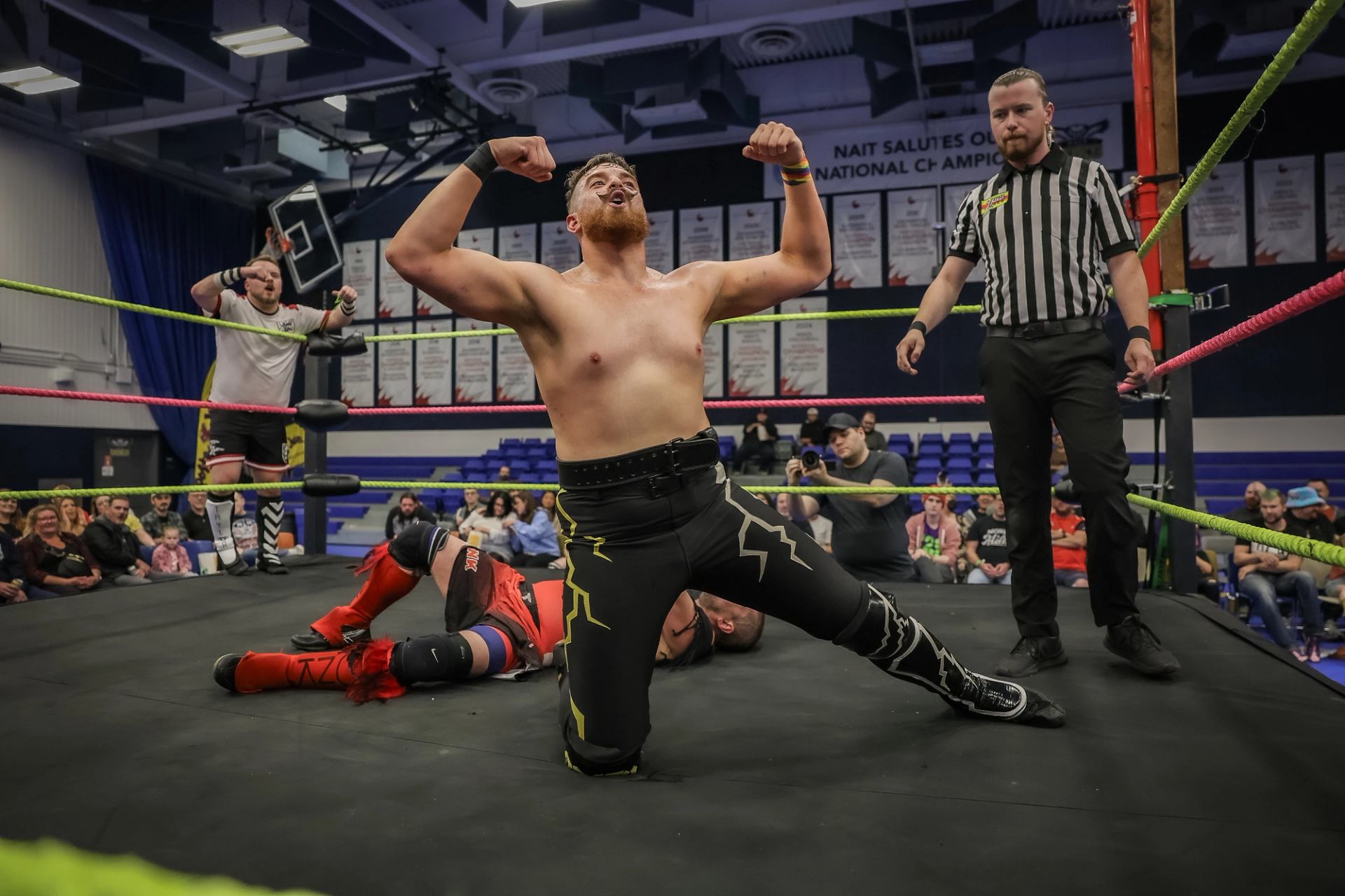 Professional wrestler kneels flexing in ring as opponent lies on mat, referee watching.