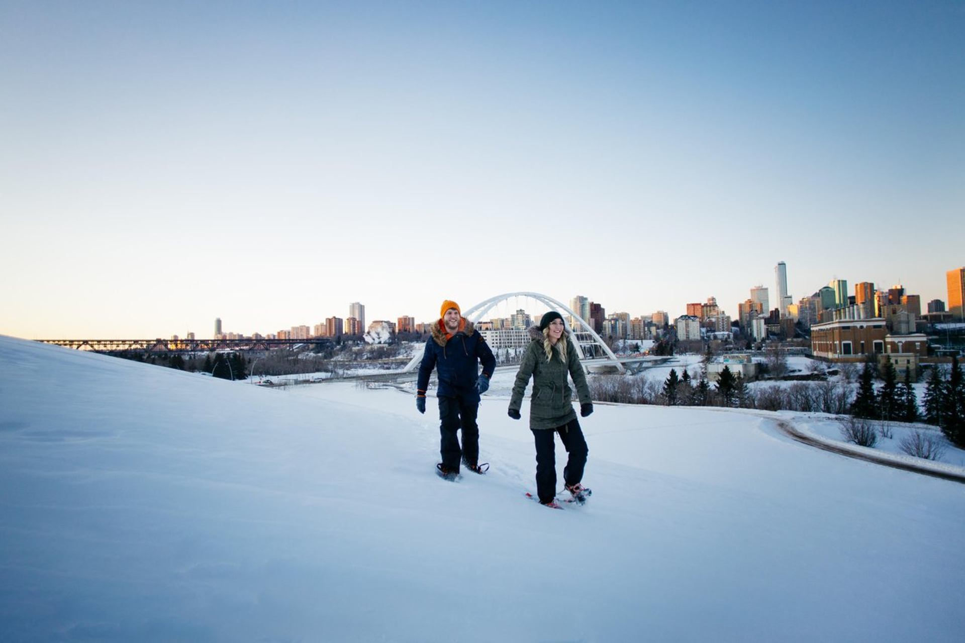 Two people snowshoeing on a snowy hill overlooking a city skyline with a bridge.