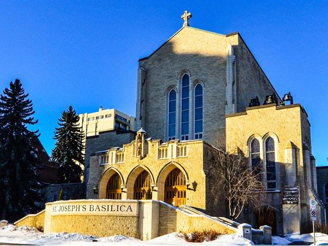 Exterior of St. Joseph’s Basilica with stone façade, arched entrance, and cross under a clear blue sky.