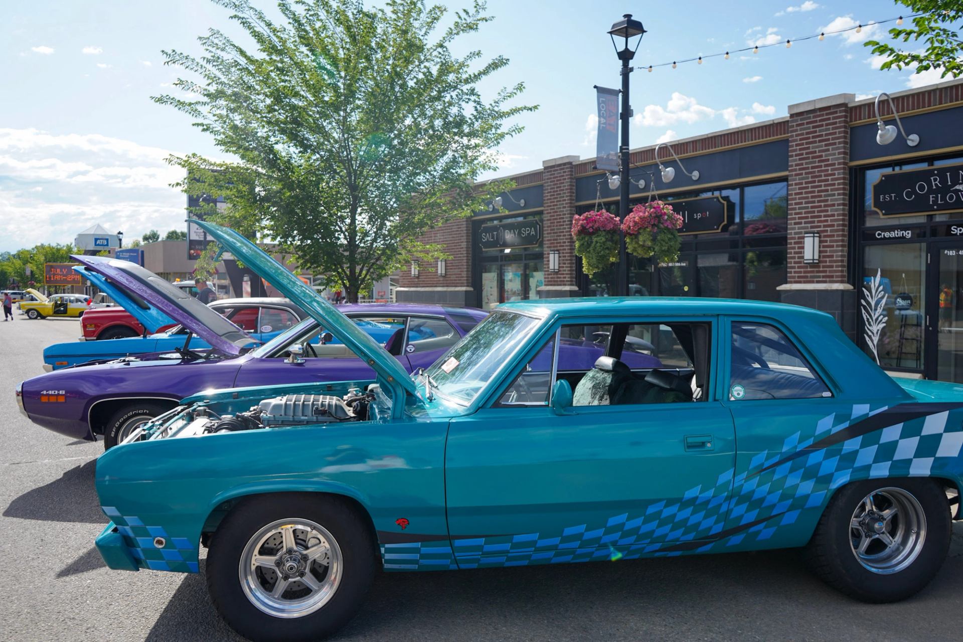 Classic teal and purple muscle cars with open hoods parked along a downtown street.