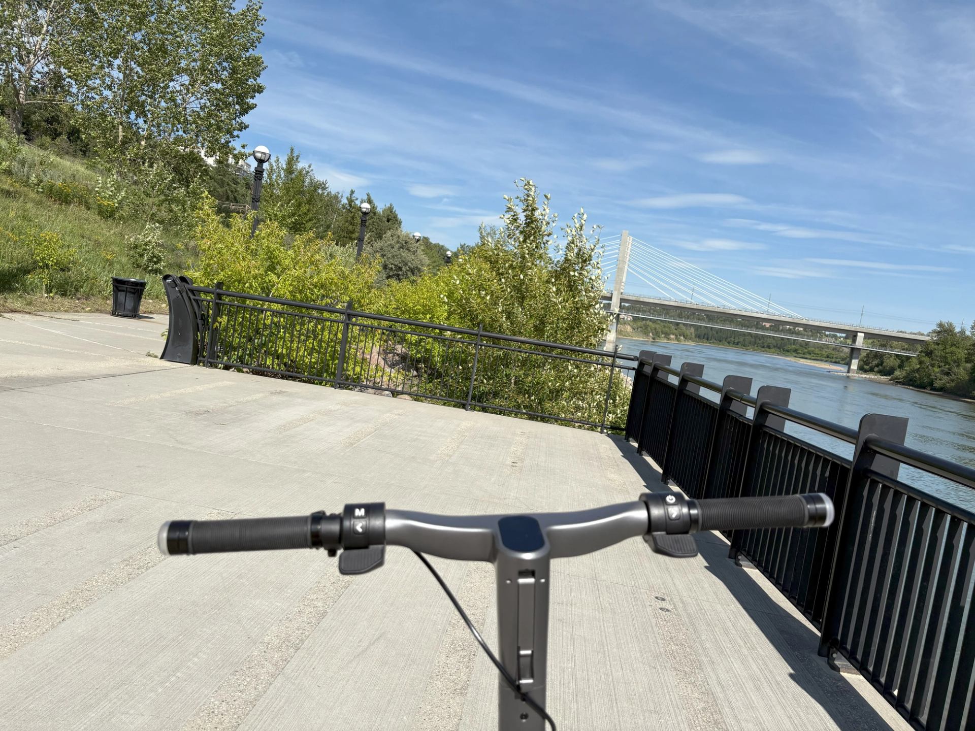 Scooter handlebars on waterfront path with trees, bridge, and clear sky in background.
