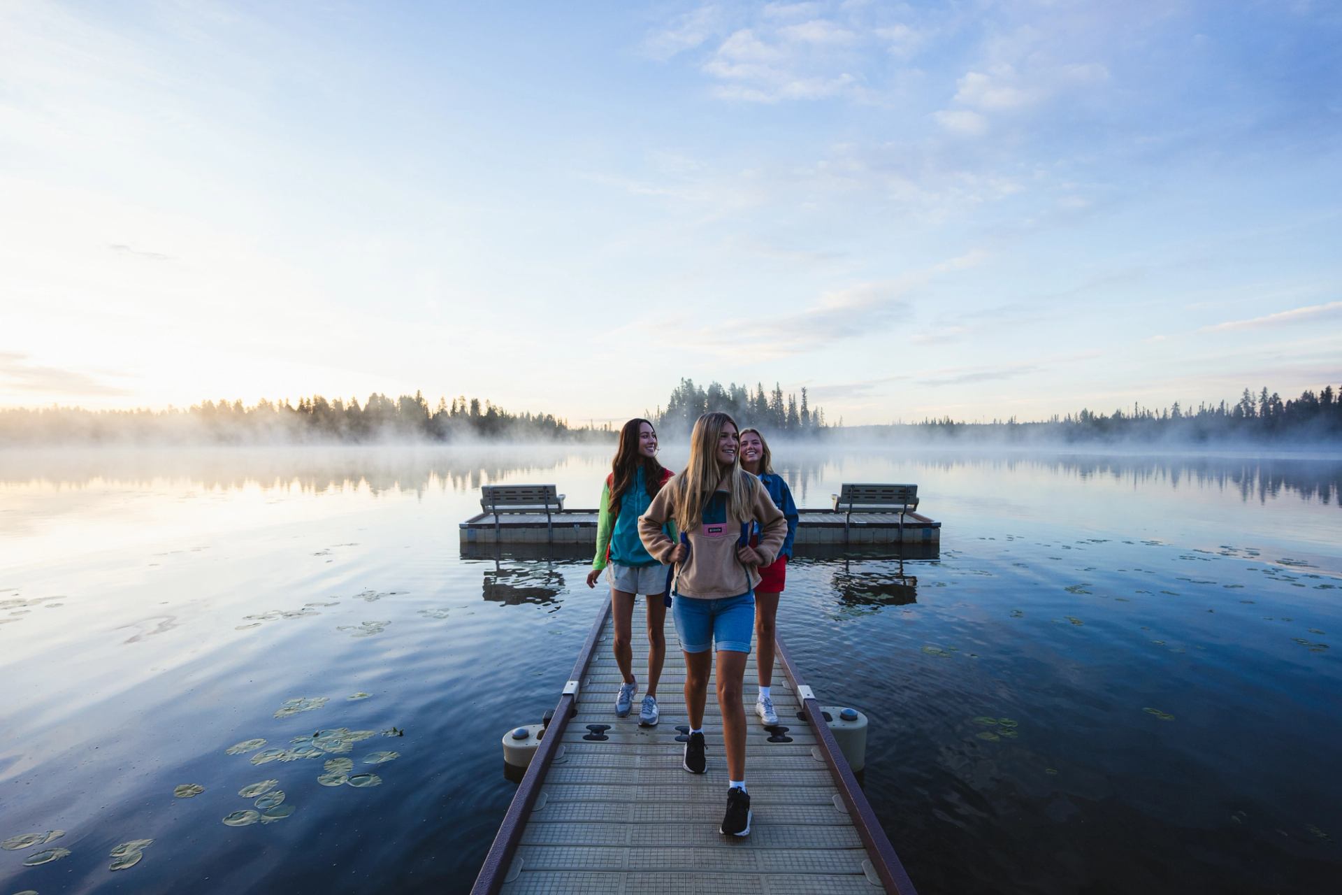 People walking on misty dock over calm lake with soft sky and trees.