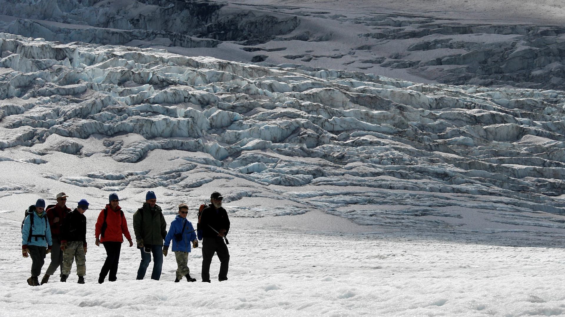 Hikers walk across a glacier below jagged blue ice ridges and crevasses.