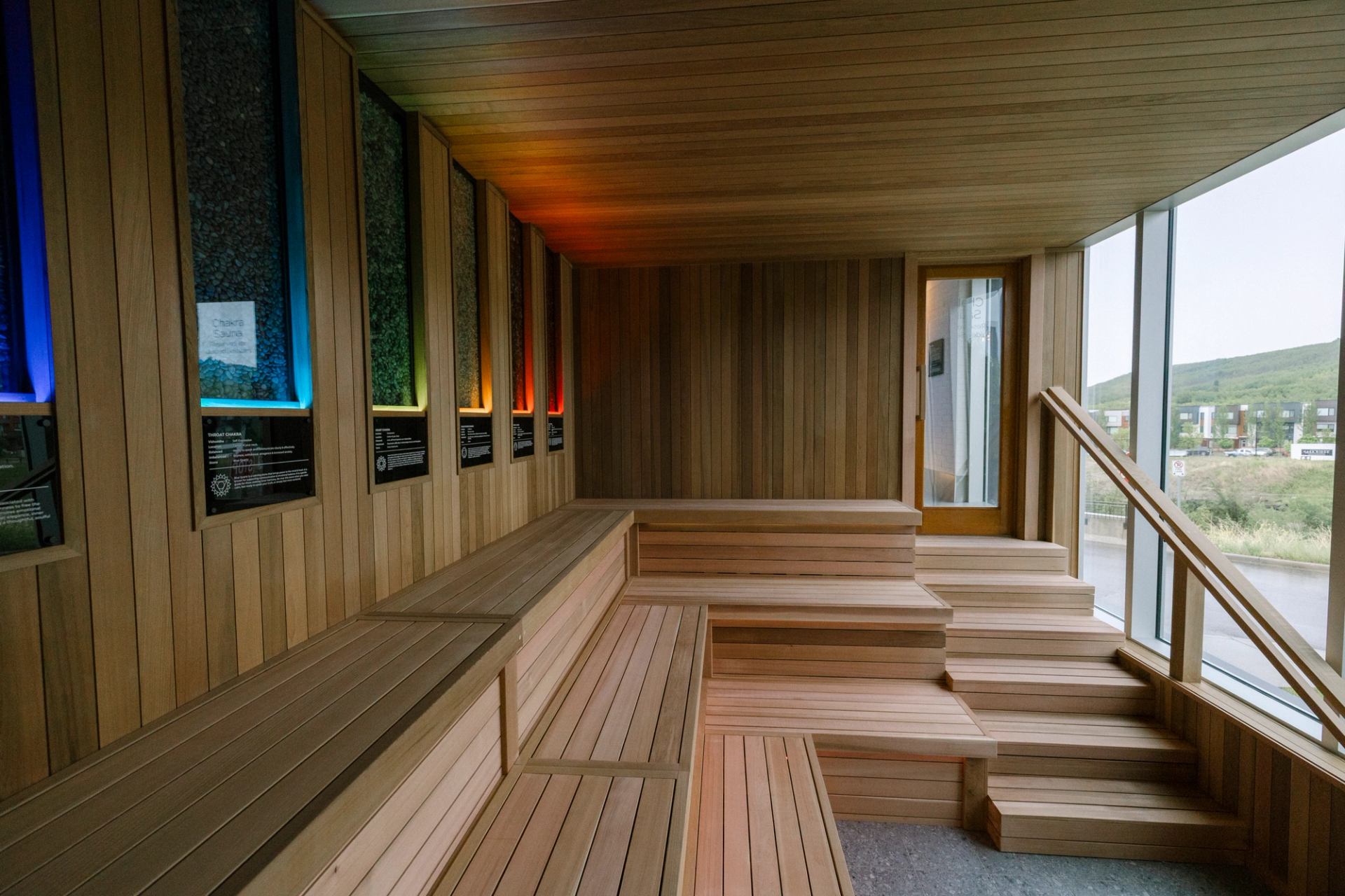 Wood-panelled sauna interior with tiered benches, coloured wall lights and large window.