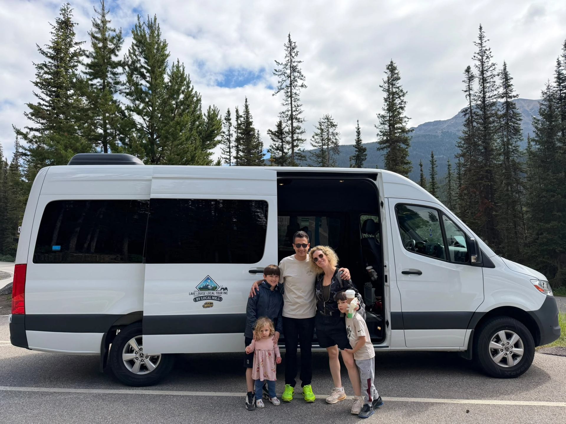 Group poses beside a white tour van in a mountain forest parking lot.