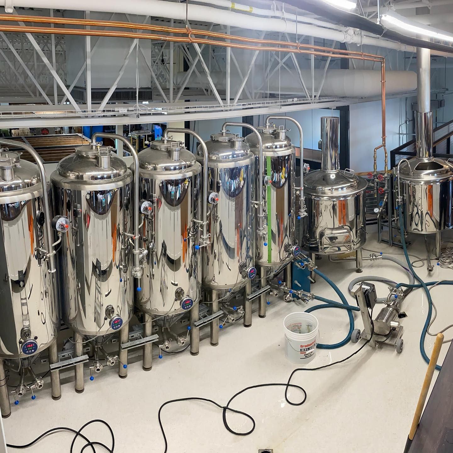 Row of shiny stainless steel brewing tanks with hoses and equipment in a brewery.