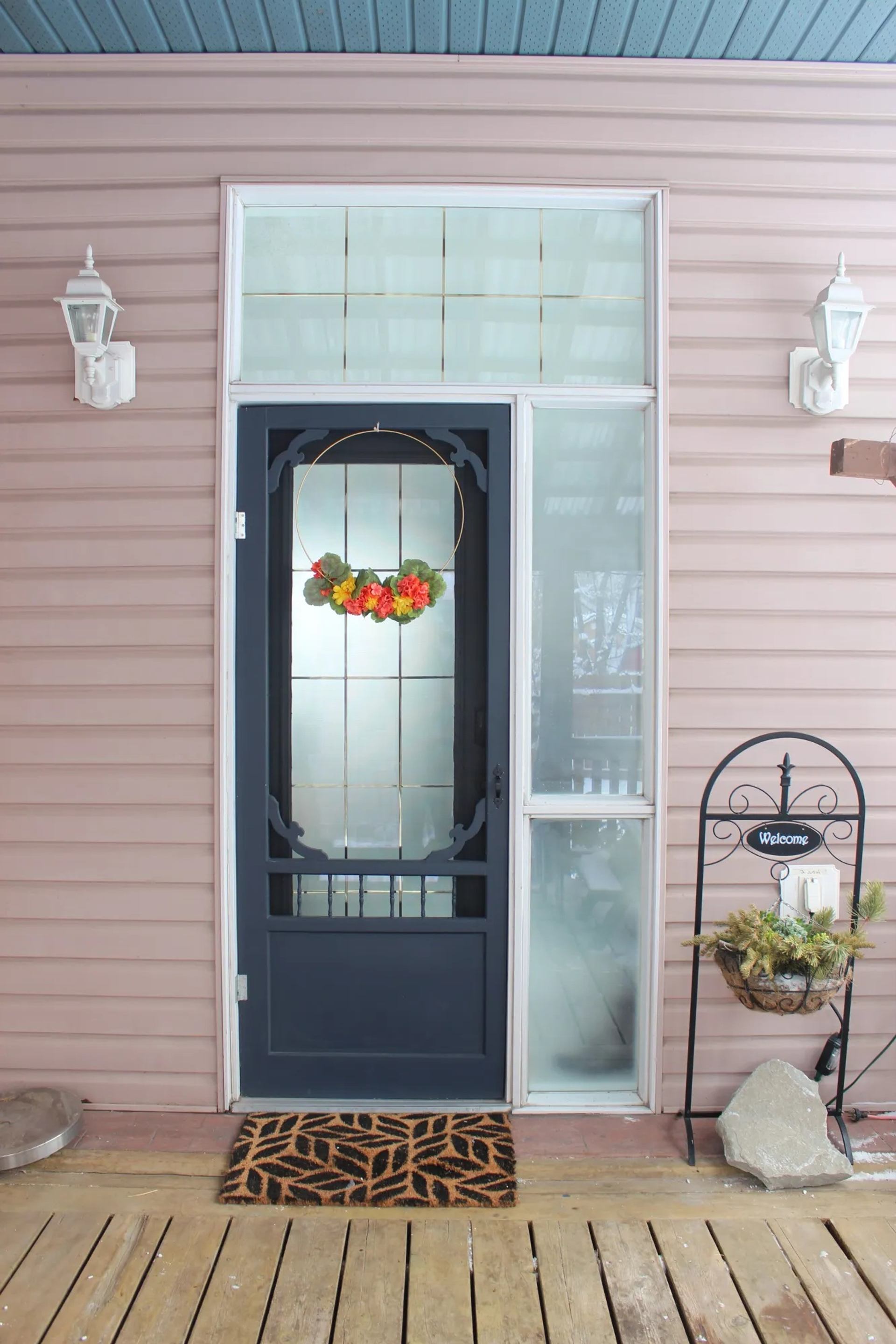 Front door with floral wreath, welcome sign, doormat and potted greenery on wooden porch.