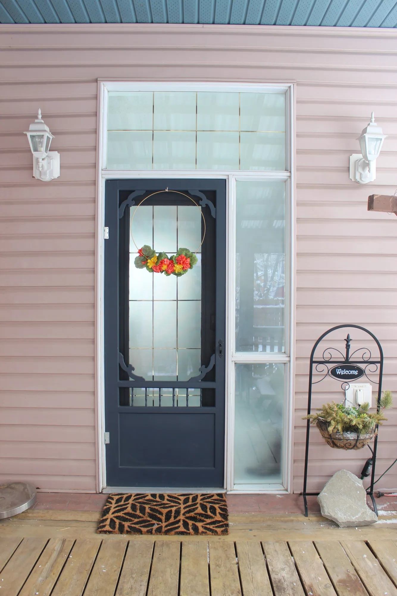 Front door with floral wreath, welcome sign, doormat and potted greenery on wooden porch.