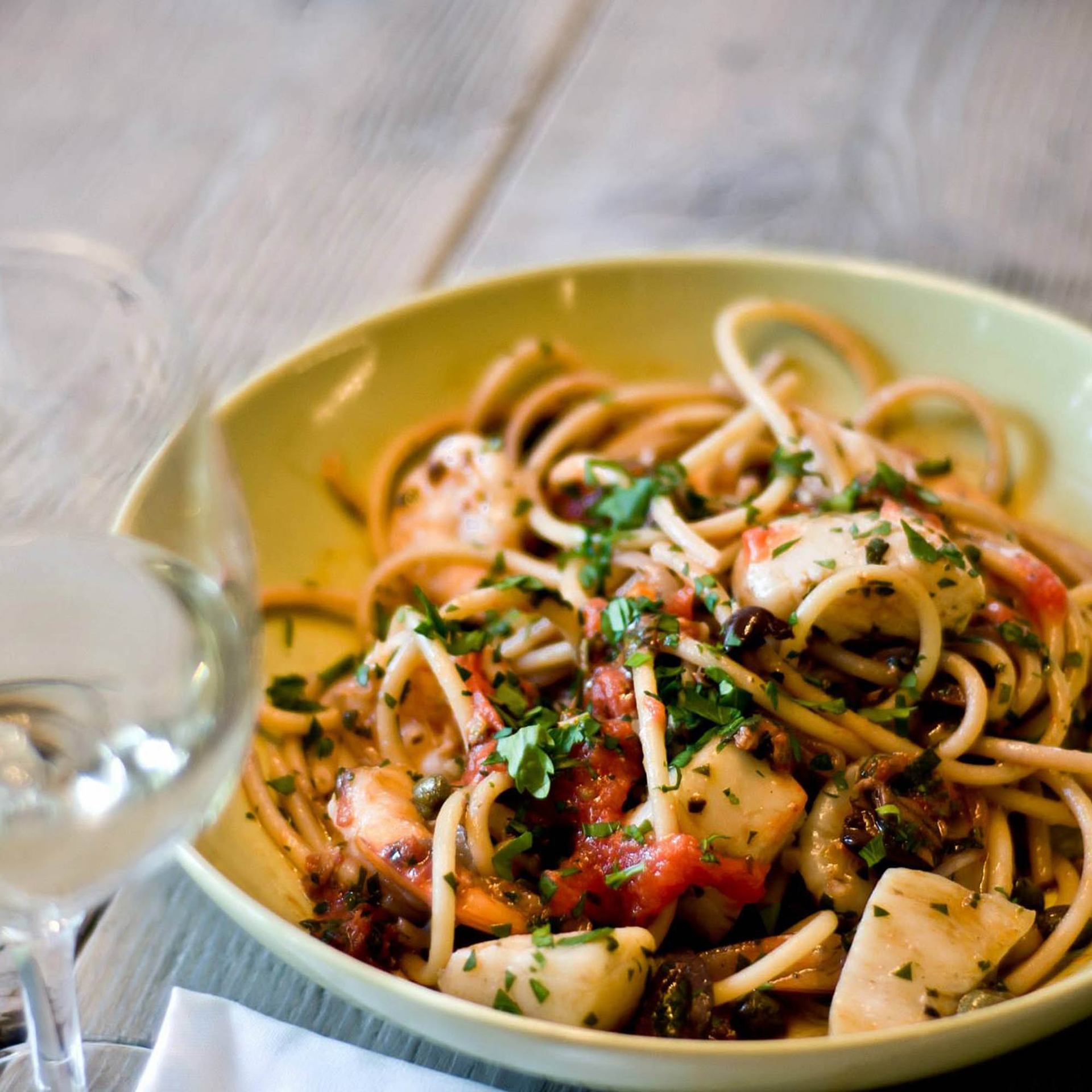 Bowl of seafood pasta with tomatoes, olives and chopped herbs beside a wine glass.
