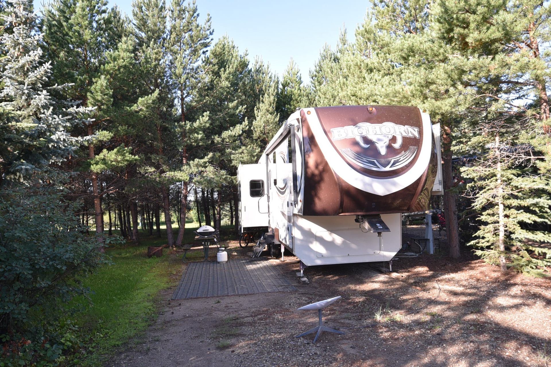 Bighorn fifth-wheel trailer at wooded RV site with picnic table and grill.