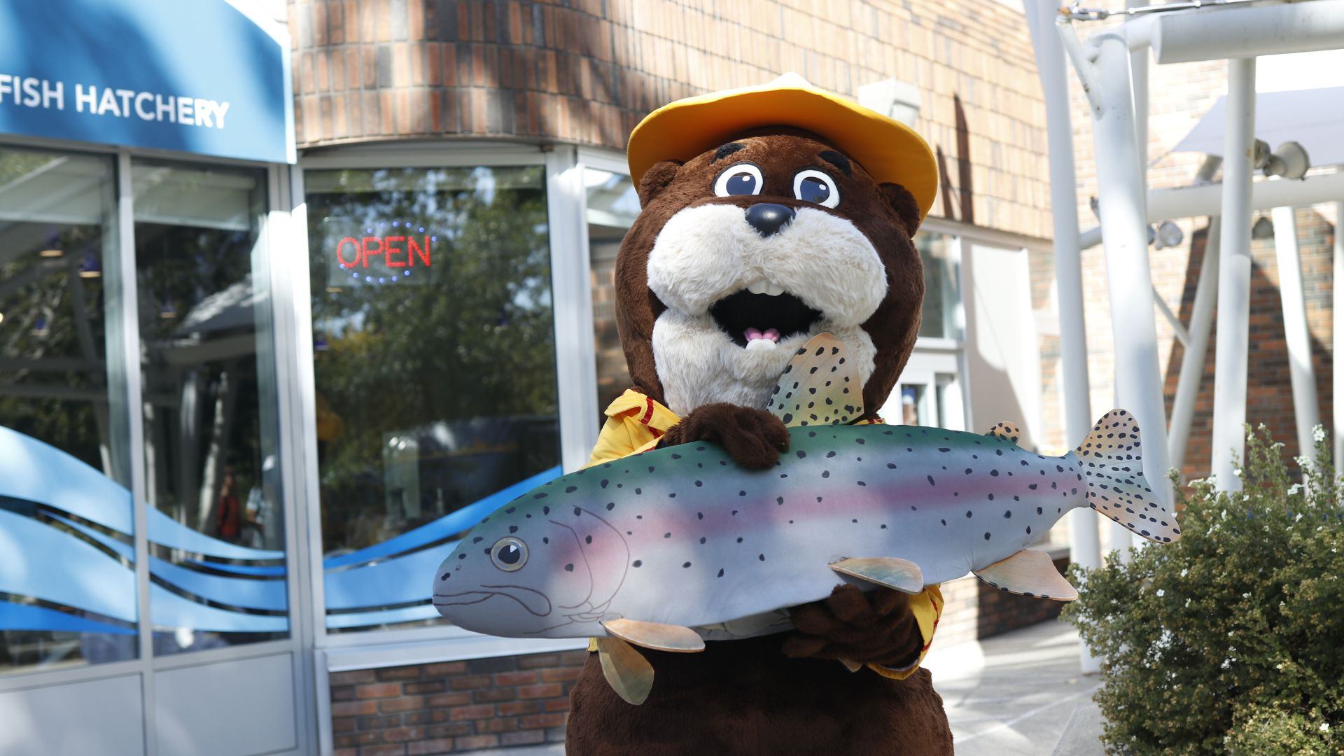 Mascot with fish model stands outside fish hatchery building.