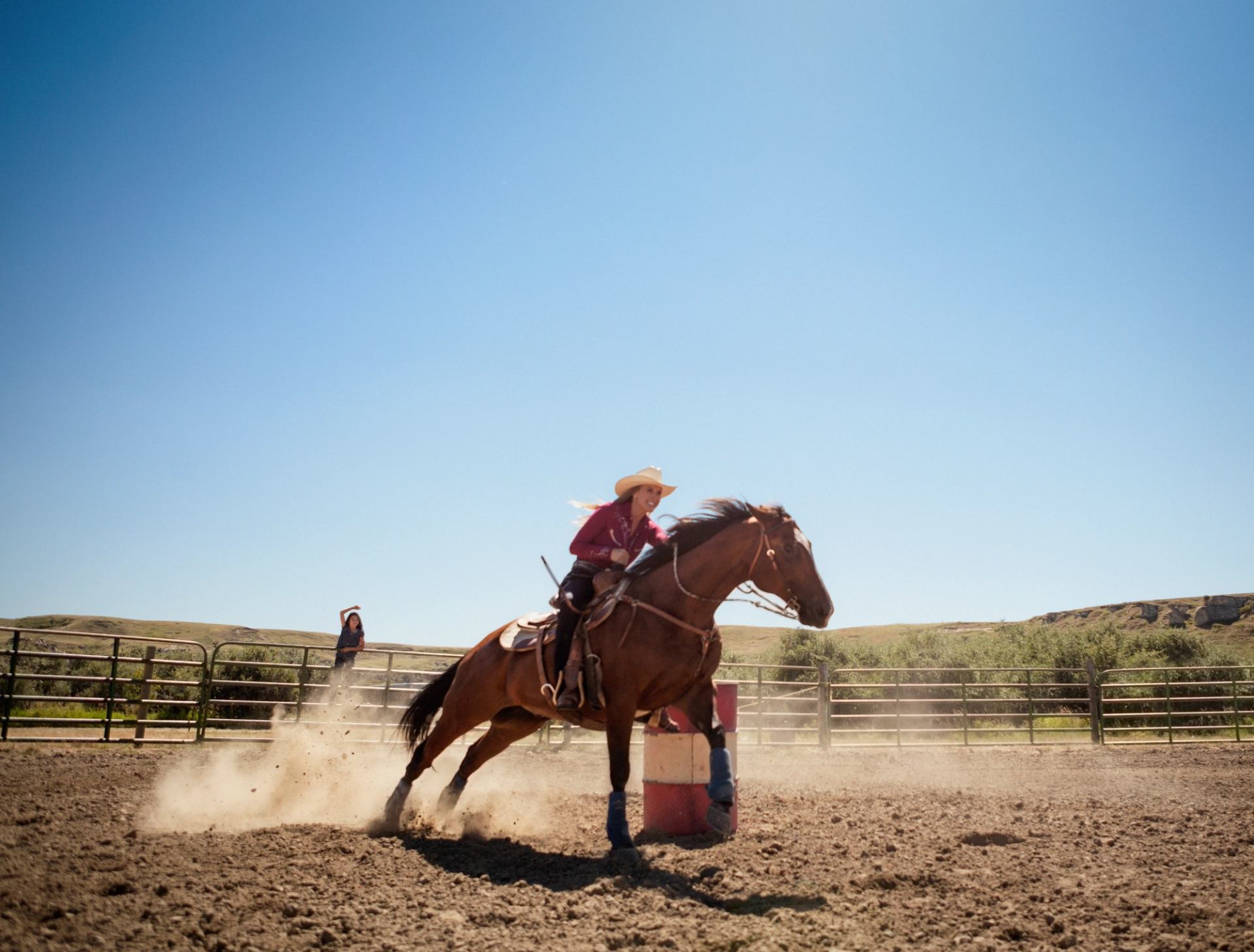 Rider on galloping horse turns around barrel in dusty outdoor arena with fencing.
