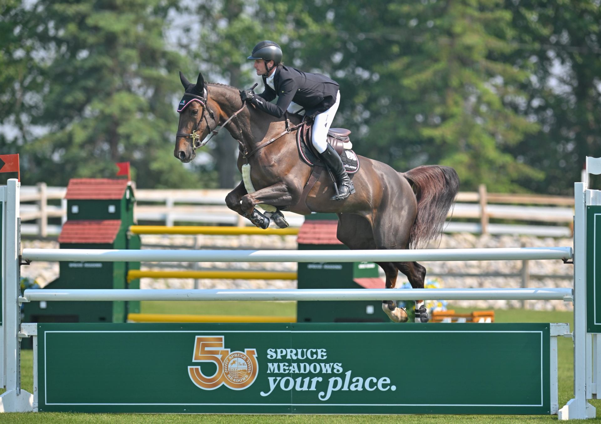 A horse and rider jump over a green fence with the Spruce Meadows logo.