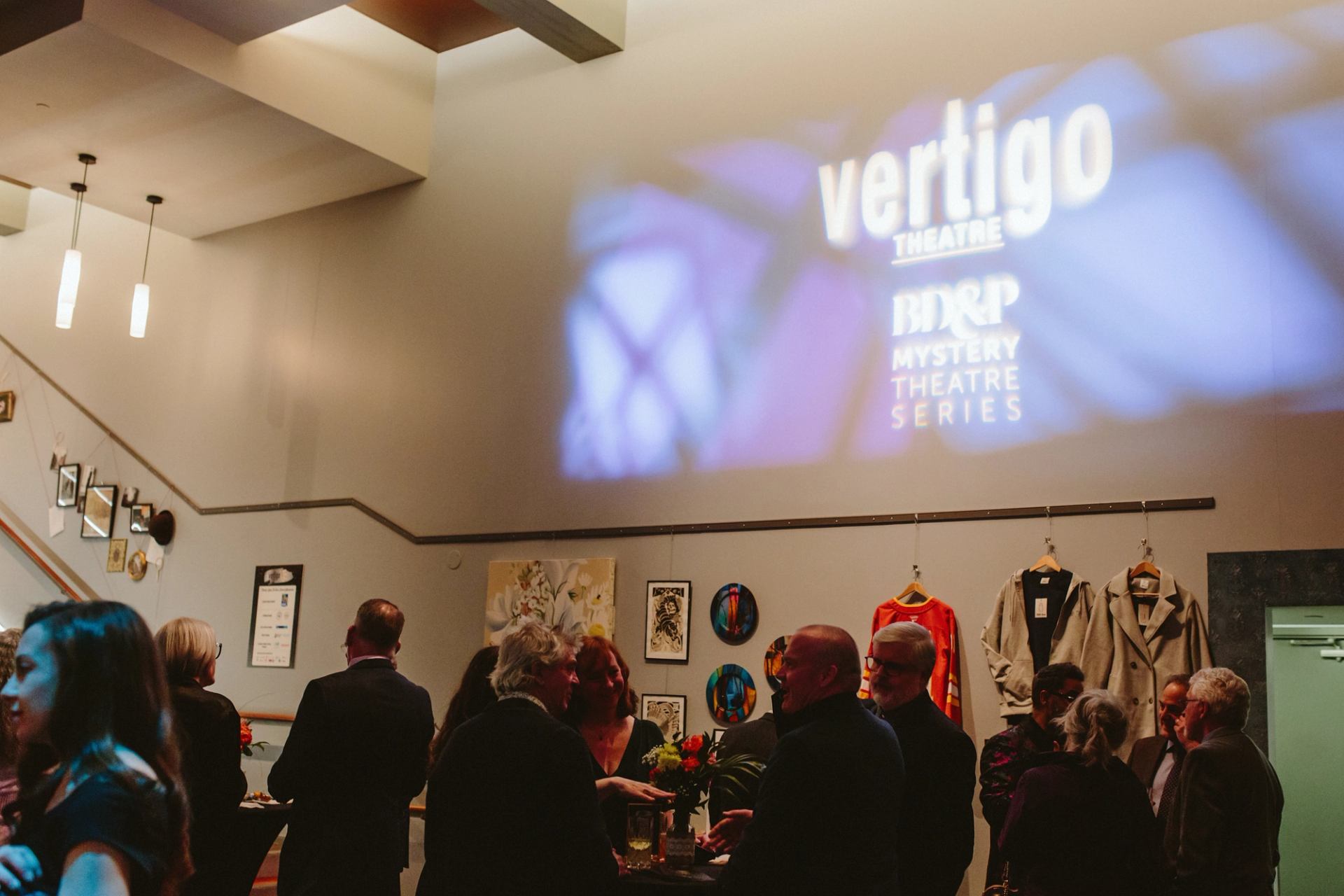 Guests mingle in theatre lobby beneath Vertigo Theatre mystery series projection.