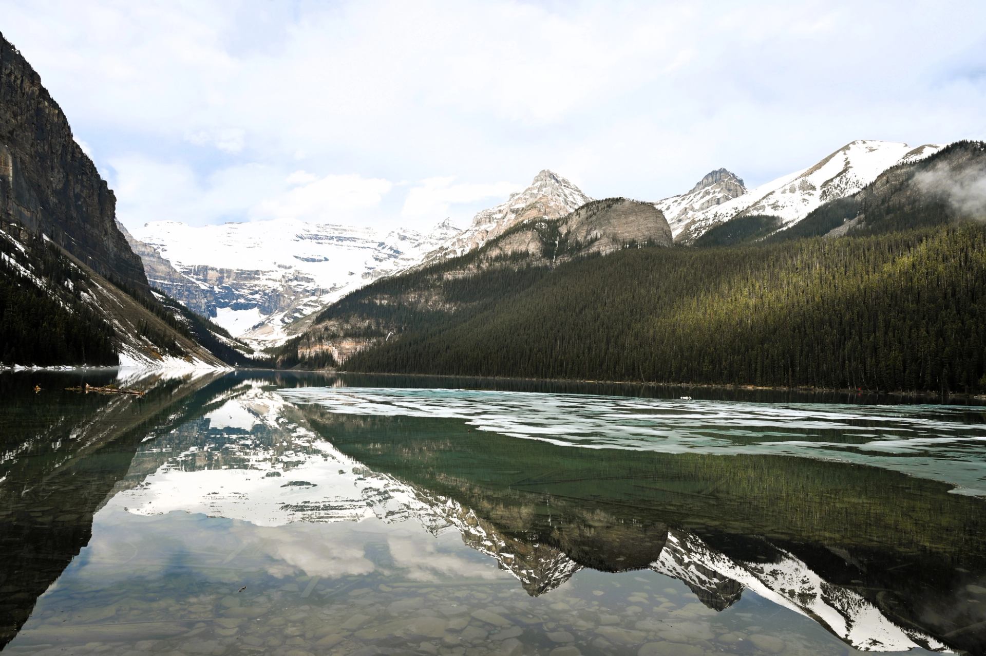 Snow-capped mountains and dense forest reflected in a calm, partially frozen lake.