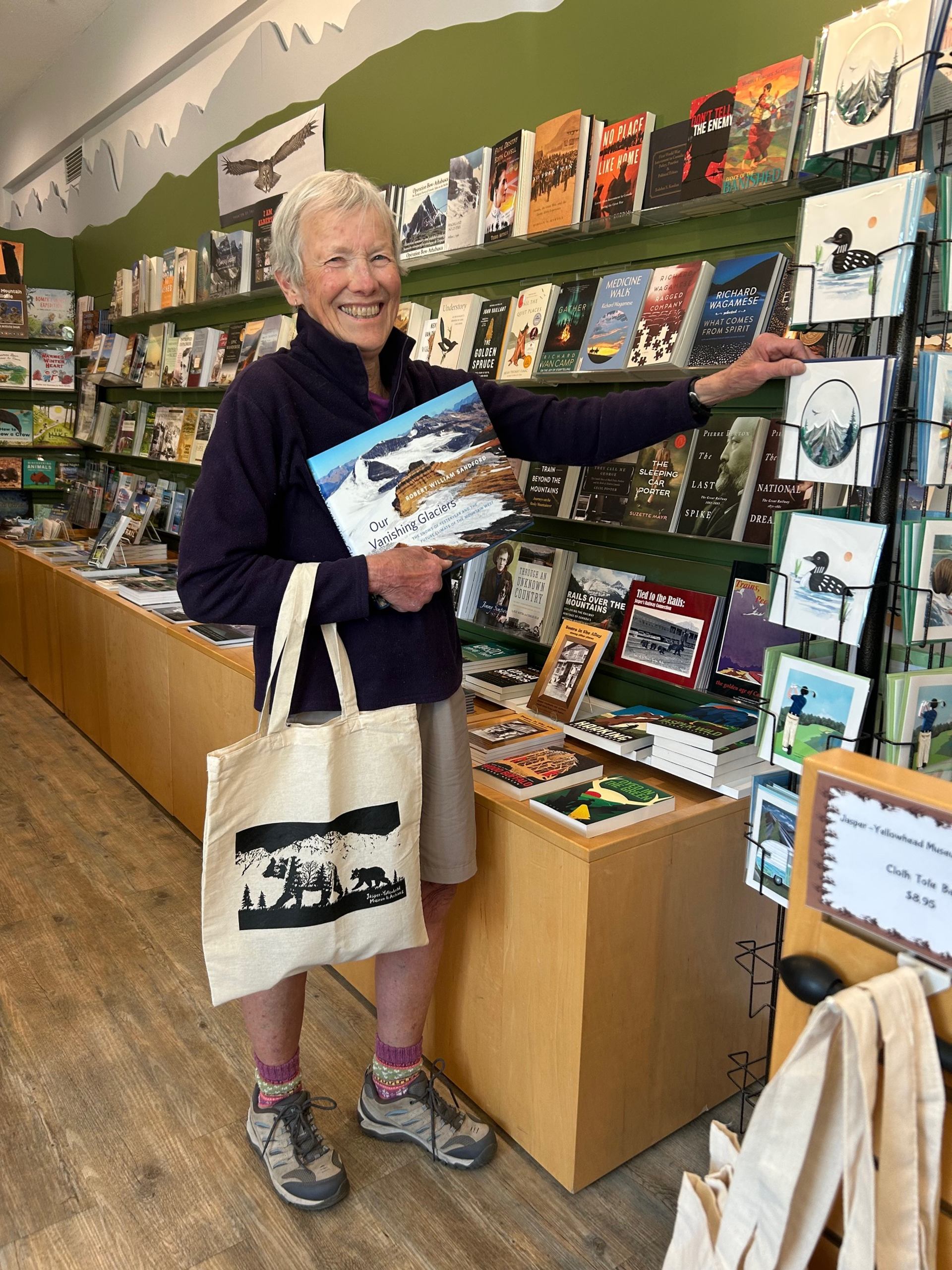 Smiling person in bookstore holding glacier book and tote bag beside postcard rack.