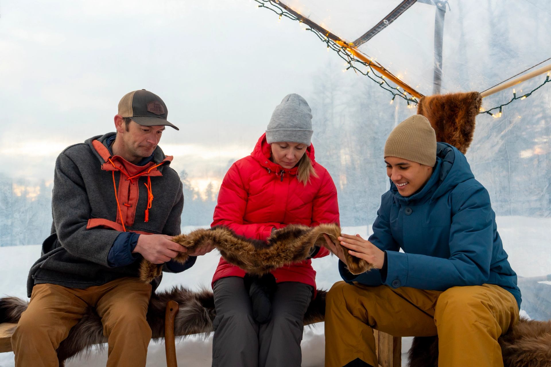 Three people seated inside a tent examining a piece of fur with snow outside