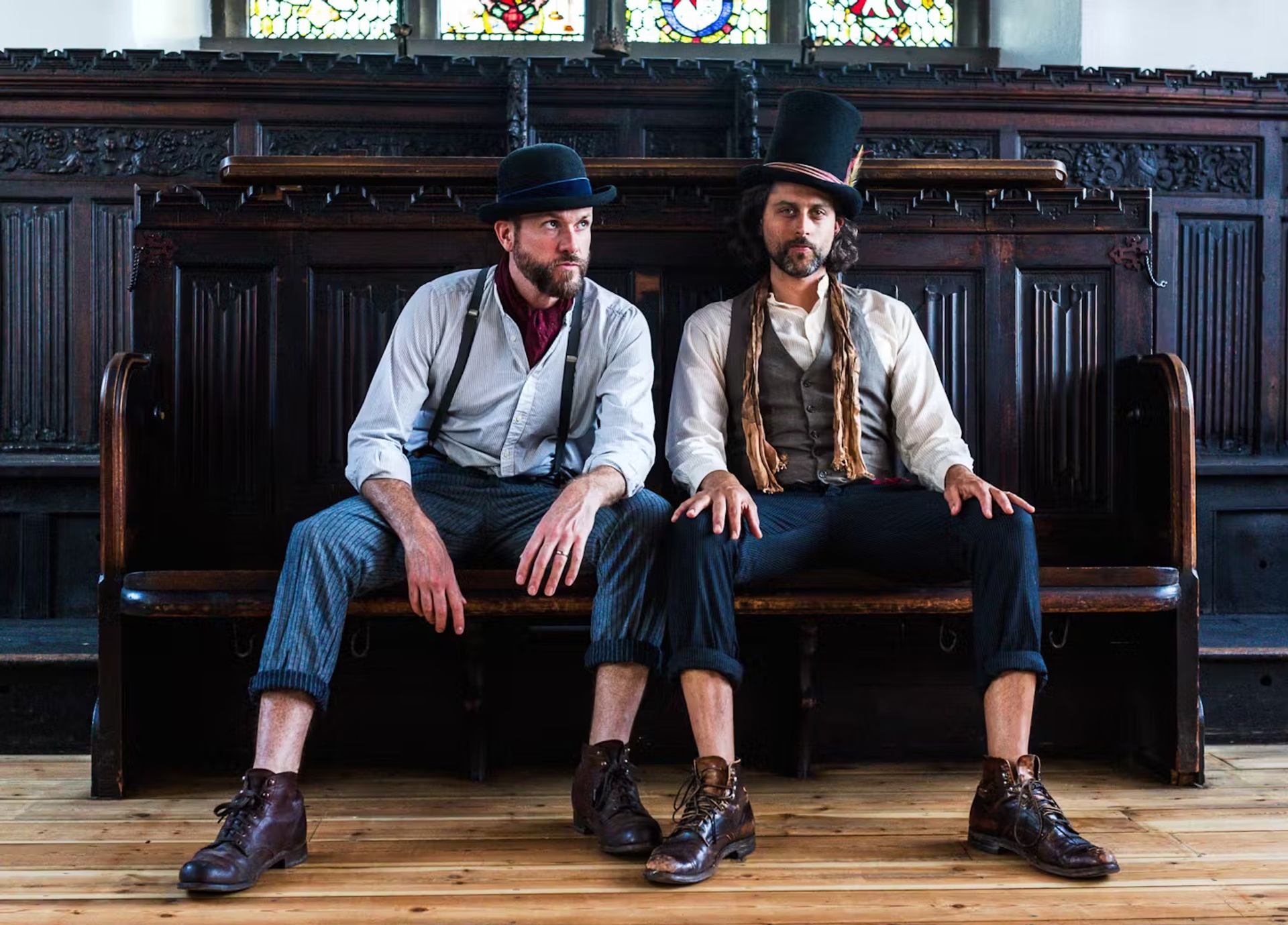 Two men in vintage clothes sit on a bench before a stained glass window.