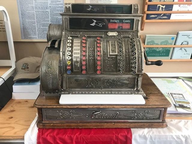 Antique metal cash register on display inside the Alix Wagon Wheel Museum with pamphlets behind it.