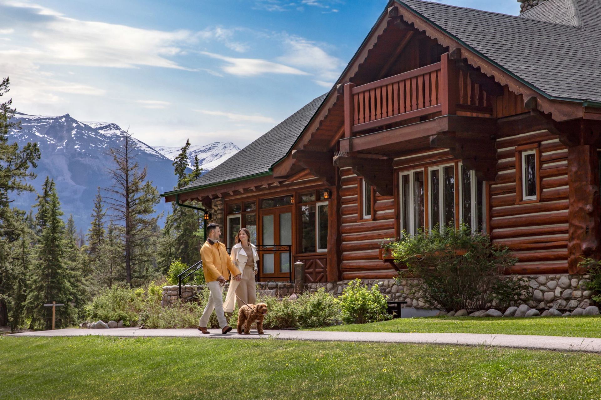 Log cabin with large windows and balcony, couple walking dog on path, mountains behind.