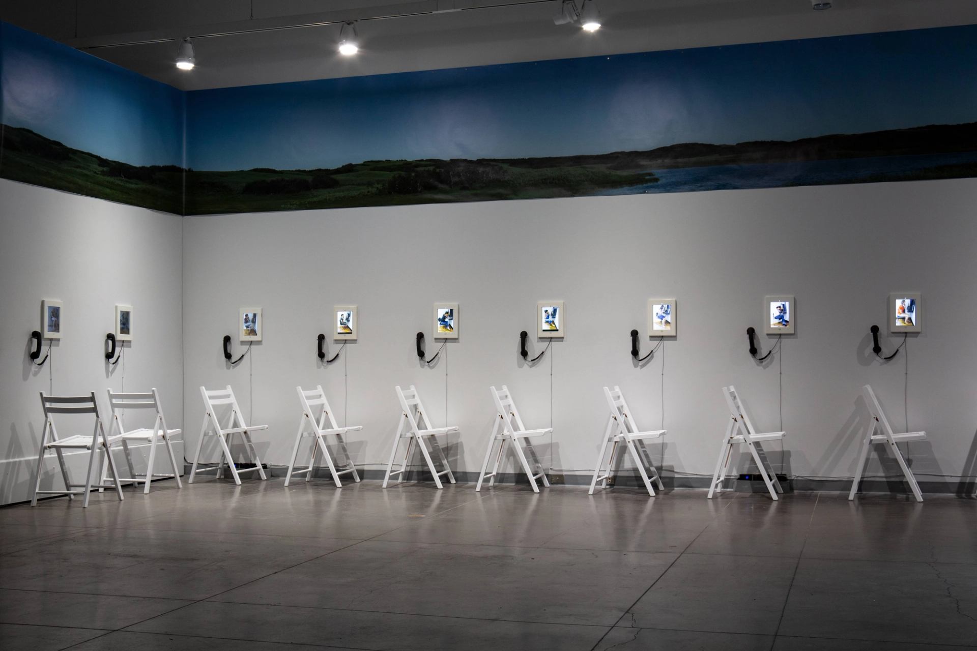 A gallery installation with a line of white chairs, small screens showing blue figures with dangling phone receivers, and a landscape mural above.