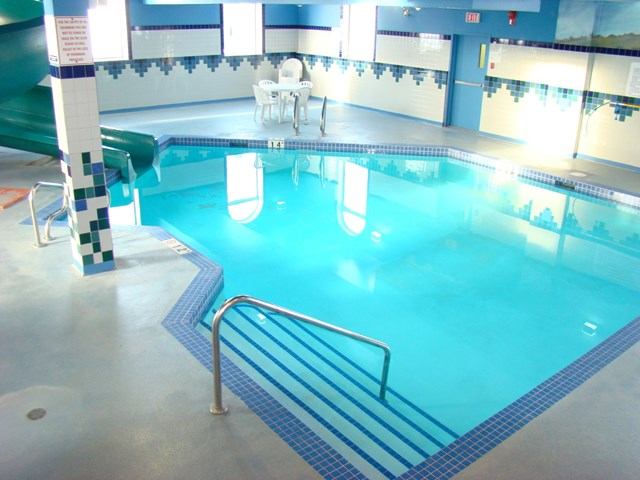 Indoor swimming pool with handrail steps, green waterslide and chairs on tiled deck.