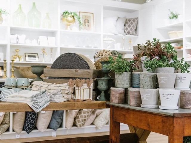 Display shelf in a home store with potted plants, folded linens, and decorative hats.