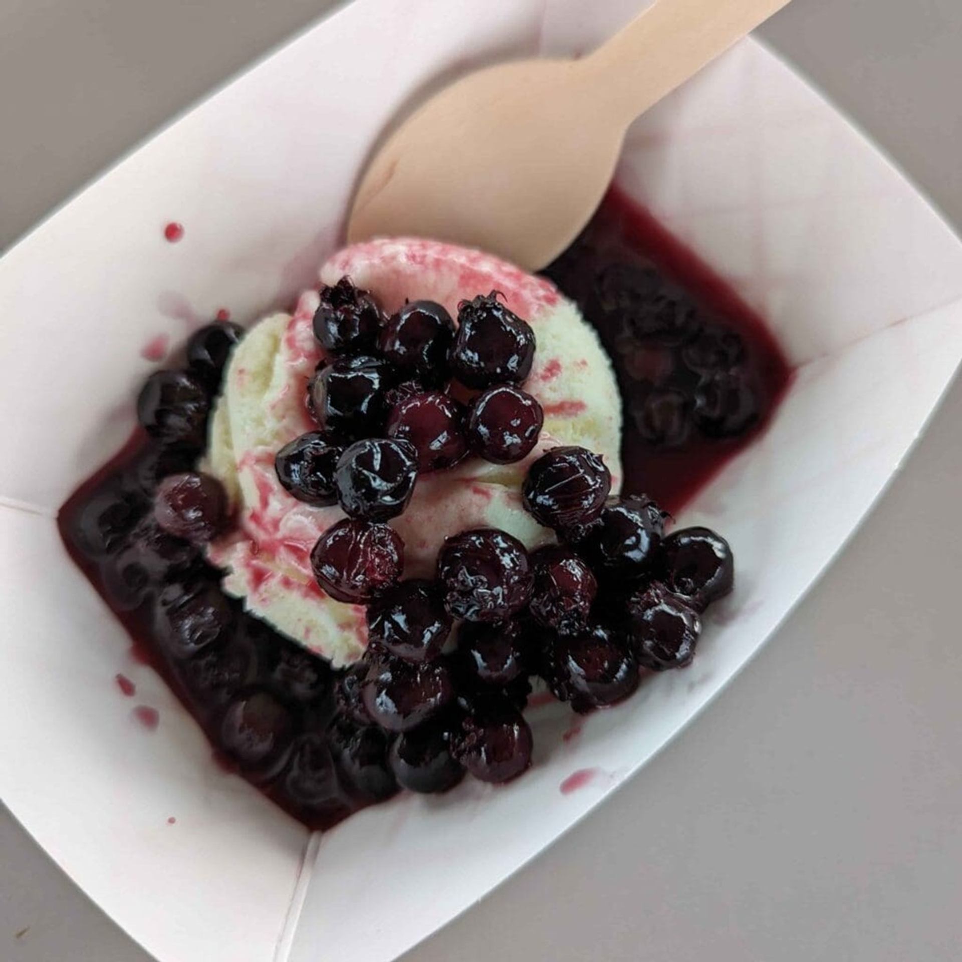 Vanilla ice cream in paper tray topped with dark berries and wooden spoon.