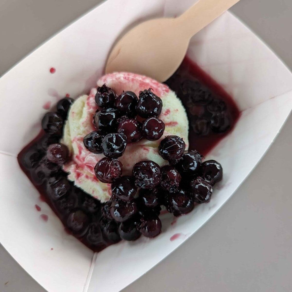 Vanilla ice cream in paper tray topped with dark berries and wooden spoon.