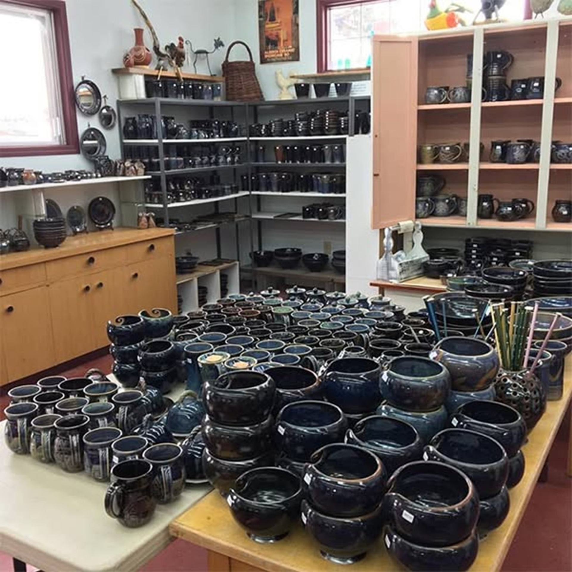 Shelves and tables filled with dark glazed pottery bowls, mugs and vases in a studio.