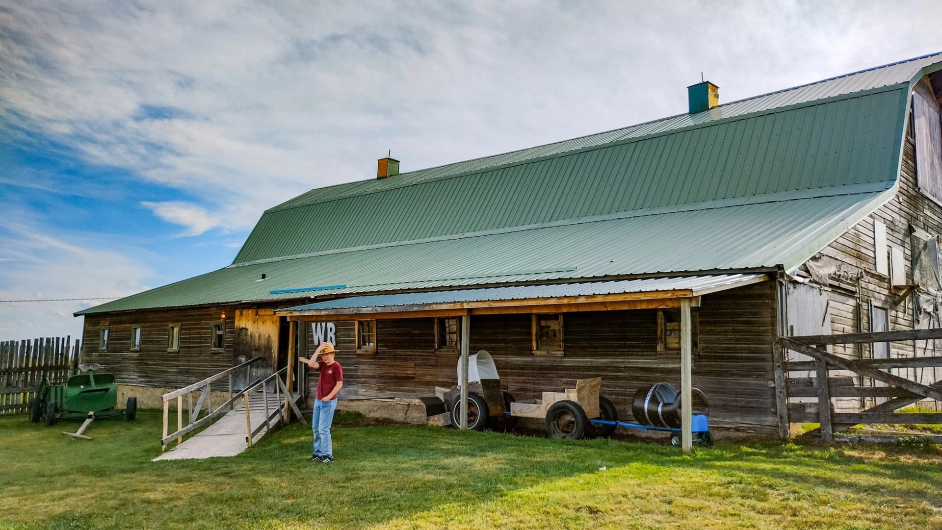 Wooden barn with green metal roof, person in cowboy hat standing on grass.