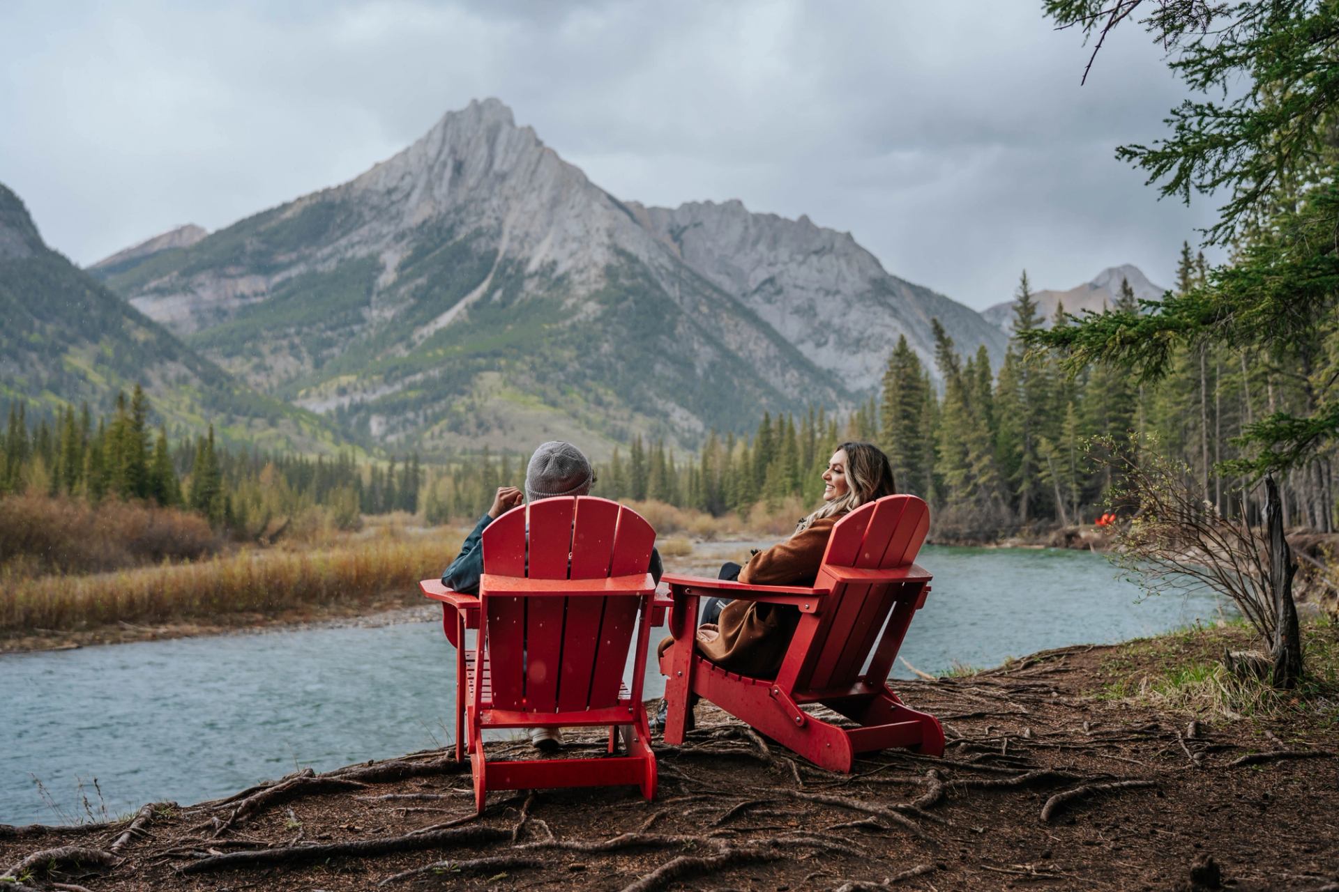 Two people sit in red chairs by a river, facing forested mountains.
