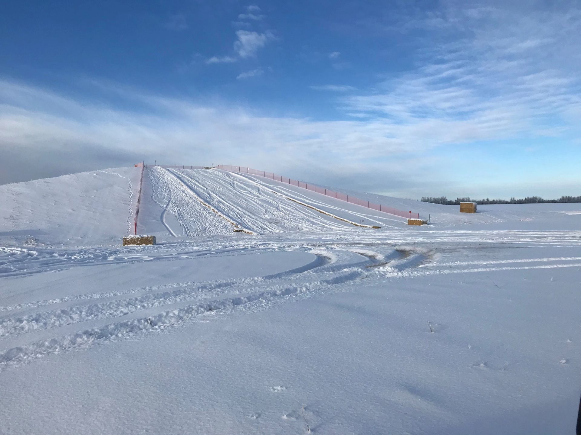 Snow-covered ski slope with sled lanes, hay bales, and a fence along the hill.