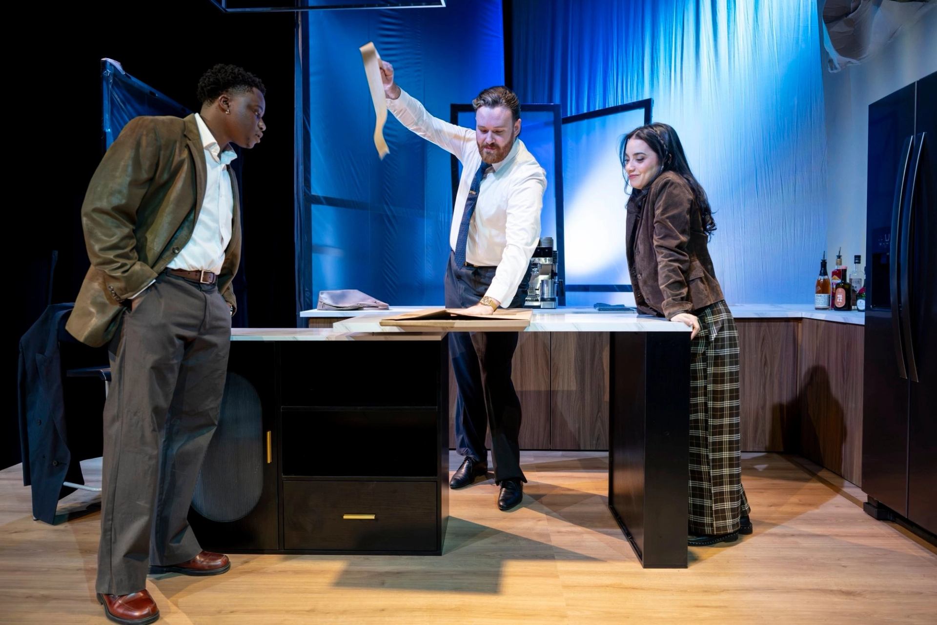 Stage scene with three people around a kitchen island examining a long sheet of paper.