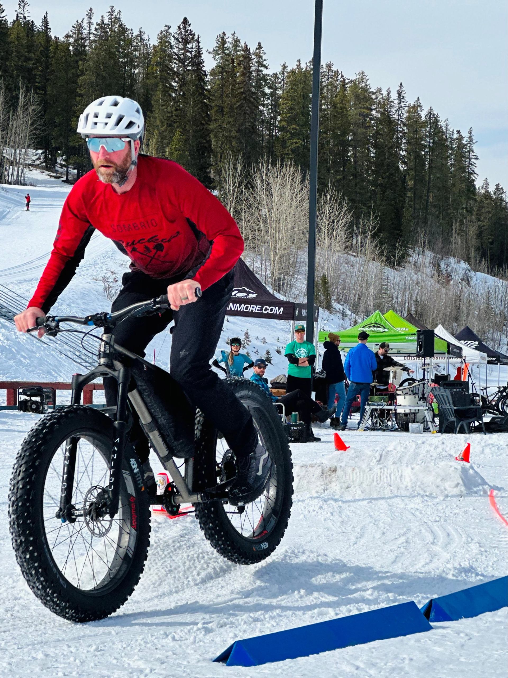 Cyclist on a fat bike navigating a snowy course with event tents and trees in the background.