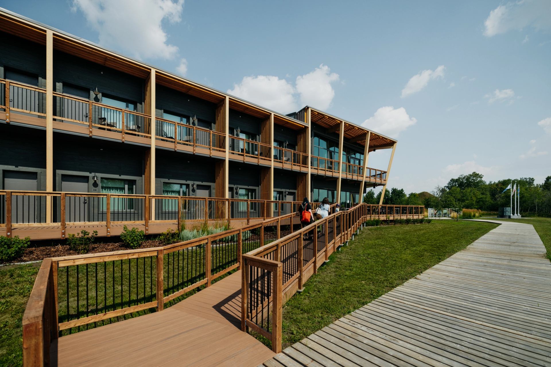Wooden two-storey lodge with balcony rooms, ramp walkway and people approaching entrance.