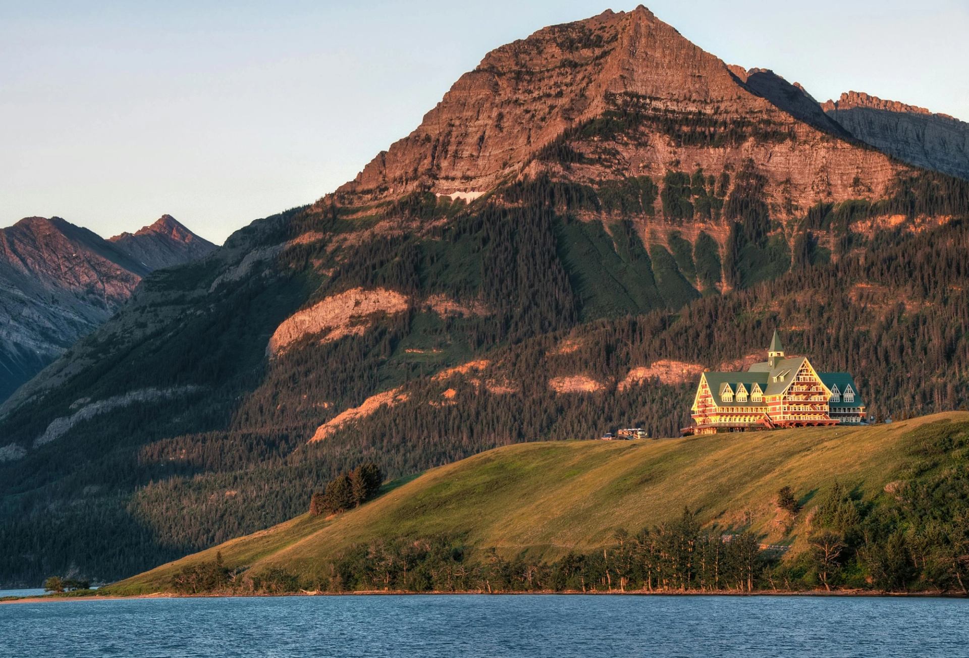 Lakeside hotel with green roof on grassy hill below steep rocky mountain slope.